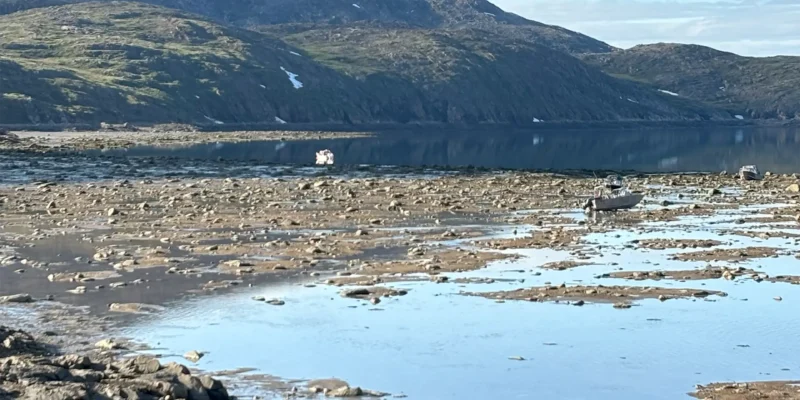 A boat floats on calm water, surrounded by rocks, with mountain rising in the background.